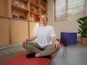 Een vrouwelijke zorgprofessional zit in een kleermakerszit op een yogamatje op de grond. Ze glimlacht en gaat beginnen aan haar yoga oefeningen.