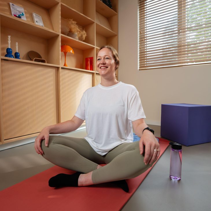 Een vrouwelijke zorgprofessional zit in een kleermakerszit op een yogamatje op de grond. Ze glimlacht en gaat beginnen aan haar yoga oefeningen.