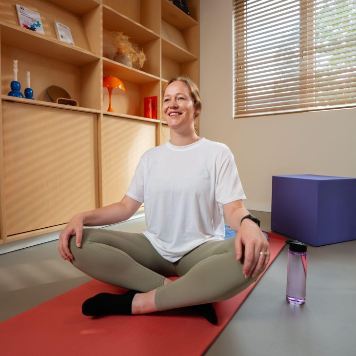 Vrouw in wit tshirt zit op oranje yoga mat in kleermakerszit