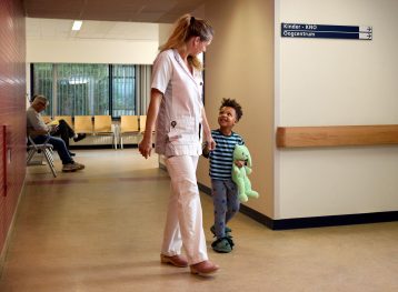 Verpleegkundige loopt op gang van ziekenhuis met een klein jongetje aan haar hand. Hij heeft een groen konijn in zijn handen en draagt een pyjama.