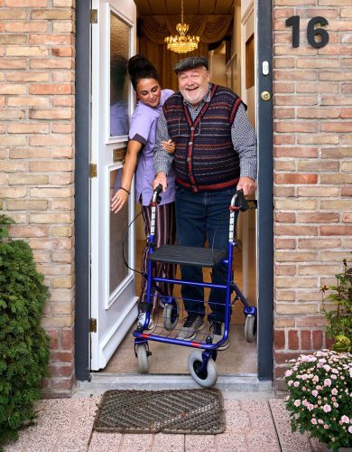 Een thuiszorgmedewerker helpt een man met rollator naar buiten.