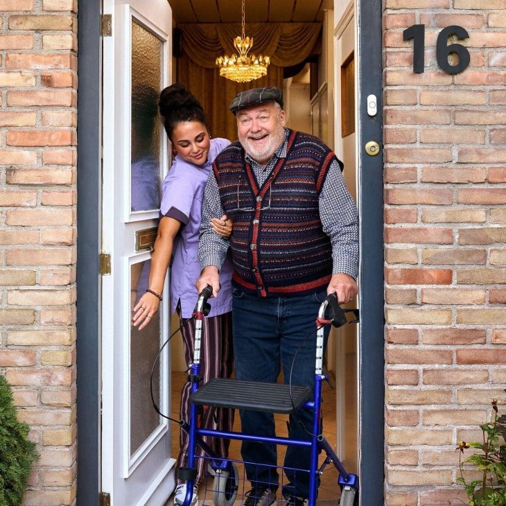 Thuiszorgmedewerker in paars pak helpt oudere man met pet en rollator door de deuropening van huis.