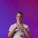 Een zorgprofessional in wit uniform maakt een hartvorm met de handen tegen een paarse en roze achtergrond