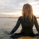 Vrouw op surfplank in het water, met wetsuit aan, bij zonsondergang
