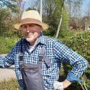Man met geblokte blouse, hoed en overal. Staat met tuingereedschap in de tuin