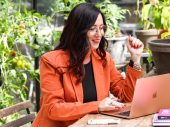 Vrouw met lang donker haar en een oranje jasje zit aan tafel met een laptop