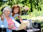 Twee vrouwen, links een oudere vrouw met een lichtblauw vestje en een laptop, rechts een vrouw met lichtbruine krullen, in natuurlijke omgeving