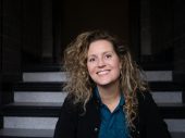Portret van vrouw met lange, lichtbruine krullen en een zwarte jas met een lichte trap op de achtergrond
