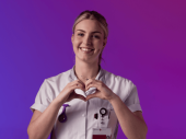 Een zorgprofessional in wit uniform maakt een hartvorm met de handen tegen een paarse en roze achtergrond.