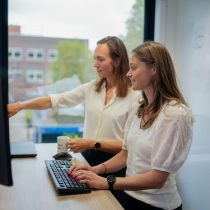 Twee vrouwen met halflang, bruin haar staan aan een bureau en wijzen naar een beeldscherm