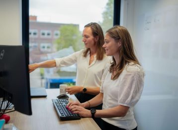 Twee vrouwen met halflang, bruin haar staan aan een bureau en wijzen naar een beeldscherm