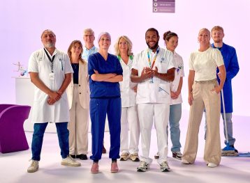 Een groep van negen zorgmedewerkers staan geposeerd voor een lichtpaarse achtergrond