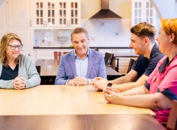 Vier mensen zittend aan een blank houten eettafel in een keuken.