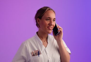 Persoon in wit uniform belt met telefoon, paarse achtergrond, pennen zichtbaar in borstzak.