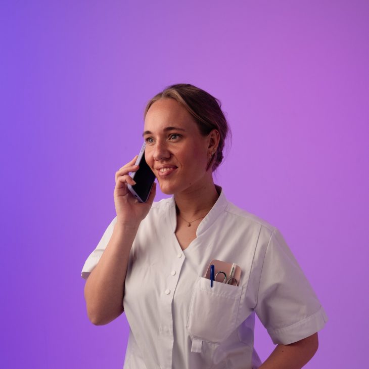 Persoon in wit uniform belt met telefoon, paarse achtergrond, pennen zichtbaar in borstzak.