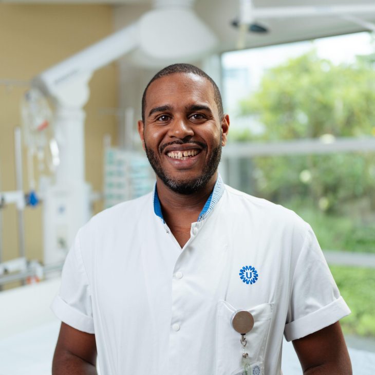 Een persoon in een wit medisch uniform staat in een ziekenhuis- of zorginstelling, met op de achtergrond medische apparatuur en een bed