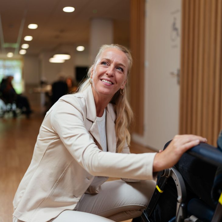 Een persoon in wit uniform biedt hulp aan iemand in een rolstoel in een moderne binnenruimte met houten vloer en sfeerverlichting