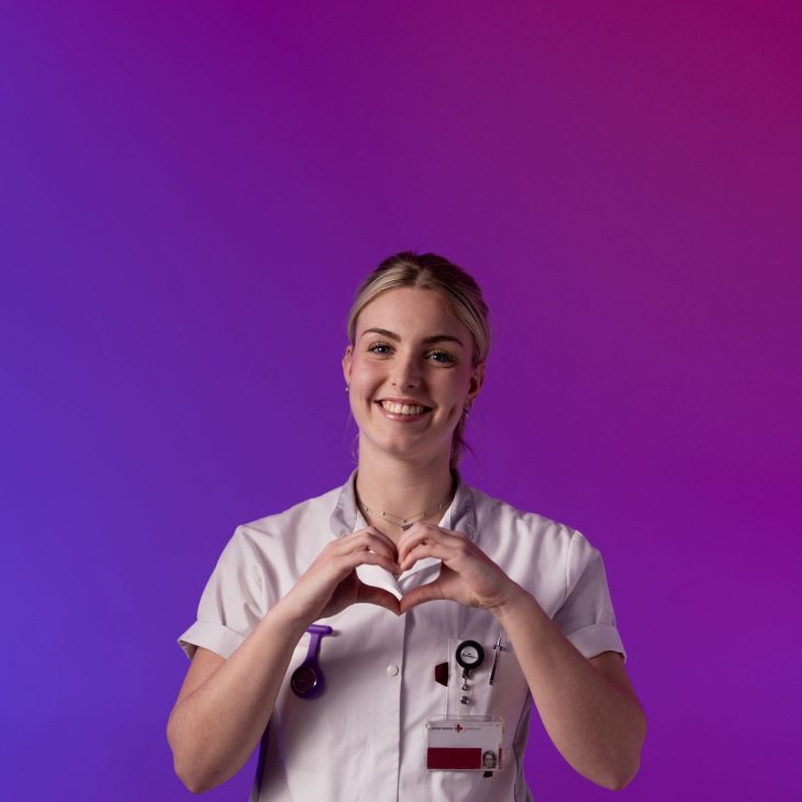 Een zorgprofessional in wit uniform maakt een hartvorm met de handen tegen een paarse en roze achtergrond