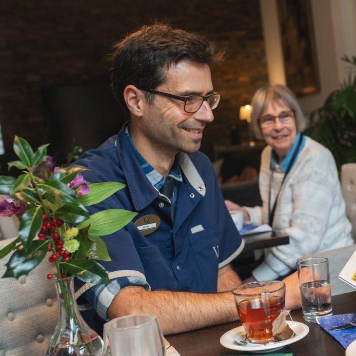 Zorgmedewerker in blauw uniform kijkt lachend naar oudere vrouw met krant
