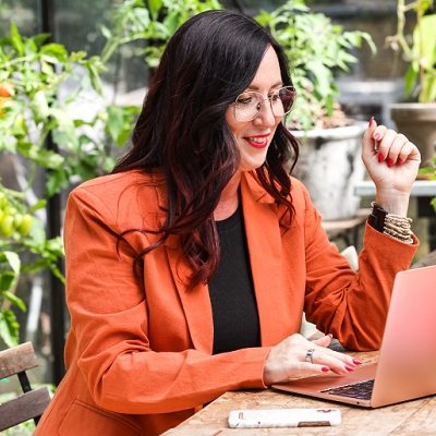 Vrouw met lang donker haar en een oranje jasje zit aan tafel met een laptop