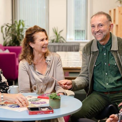 Vier mensen rondom een lichtblauwe koffietafel, lachen naar elkaar