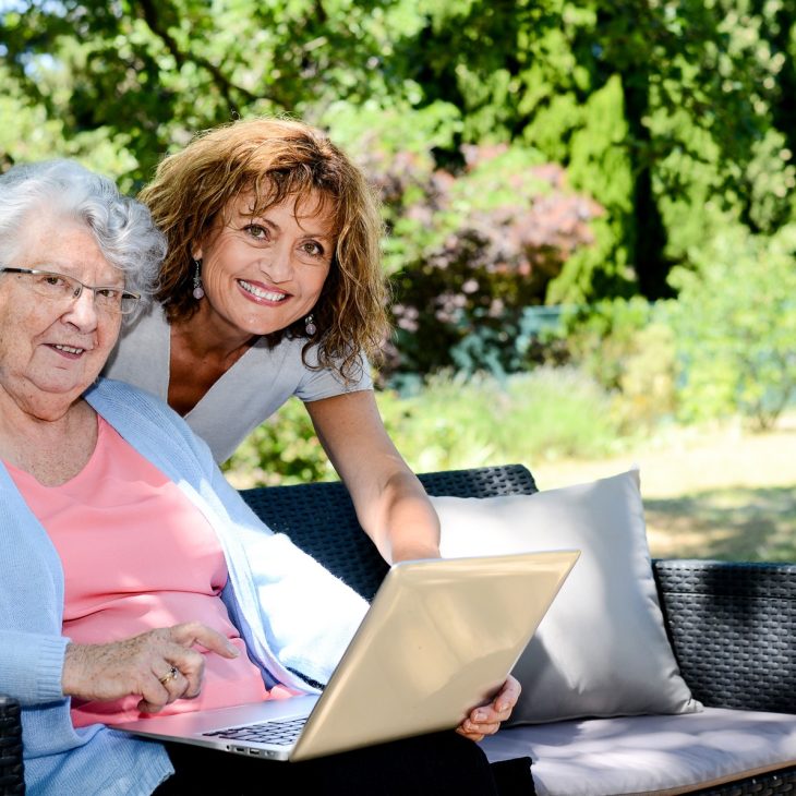 Twee vrouwen, links een oudere vrouw met een lichtblauw vestje en een laptop, rechts een vrouw met lichtbruine krullen, in natuurlijke omgeving