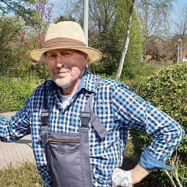 Man met geblokte blouse, hoed en overal. Staat met tuingereedschap in de tuin