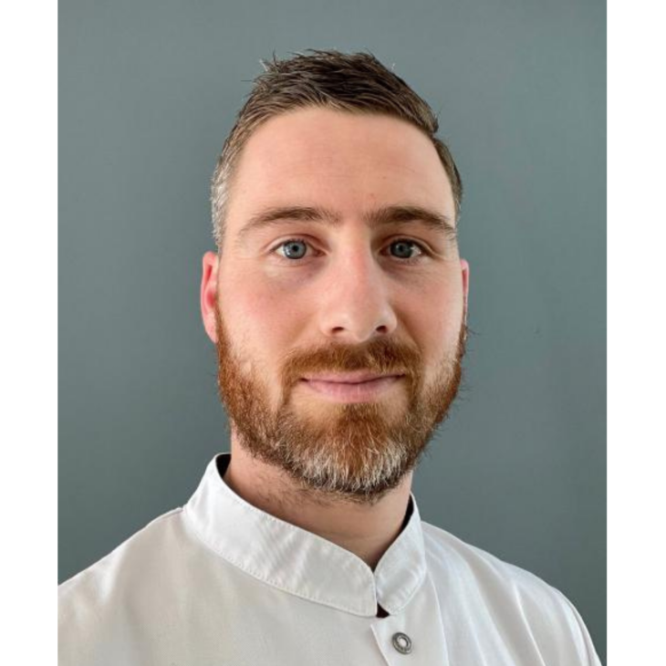 Gezicht van een man met kort haar en een bruine baard in een witte blouse, voor een grijze achtergrond