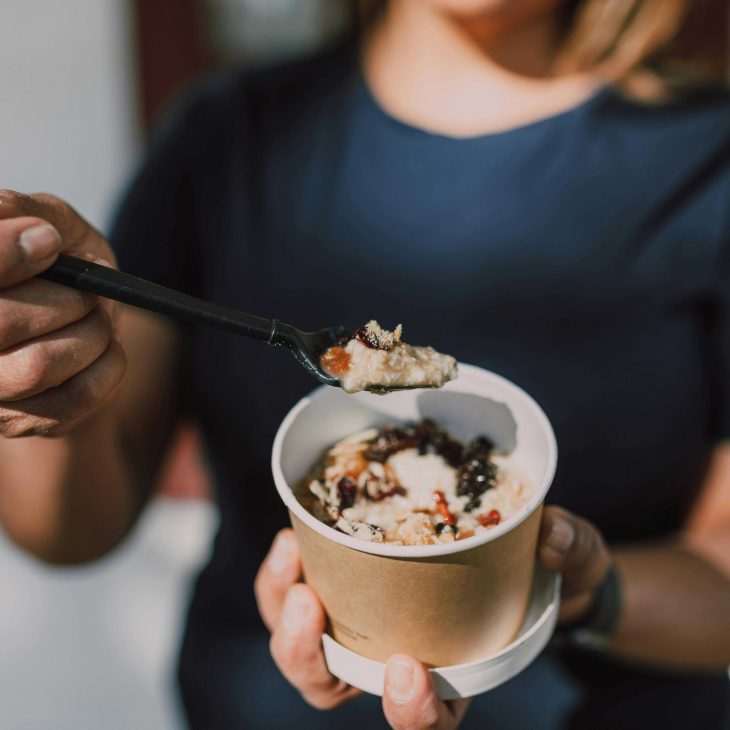 Een persoon in een donkerblauw shirt houdt een beker met dessert of snack vast en schept met een zwarte plastic lepel, mogelijk ijs of yoghurt met toppings.