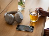 Een grijze koptelefoon ligt op een houten tafel naast een smartphone en een kop dampende thee.