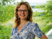 Anneke Valk, met lichtbruin haar, een blauw-witte jurk en een bril, lacht terwijl ze met haar hand in haar zij staat.