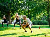 In een zonnig park doet een man een squat, terwijl op de achtergrond twee andere mensen ook aan het squatten zijn. Het gras is groen en de lente is duidelijk voelbaar.