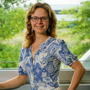 Anneke Valk, met lichtbruin haar, een blauw-witte jurk en een bril, lacht terwijl ze met haar hand in haar zij staat.