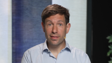 Een man met donker haar staat achter een tafel op de achtergrond zie je een plant.