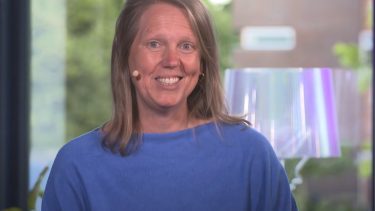 Een vrouw met een blauwe trui aan kijkt recht in de camera en heeft een glimlach, zij draagt een microfoon.