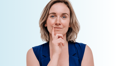 Een vrouw met een donkerblauw gilet aan die slim voor zich uit kijkt. Ze heeft haar handen over elkaar heen geslagen en één hand bij haar mond alsof ze een idee heeft.