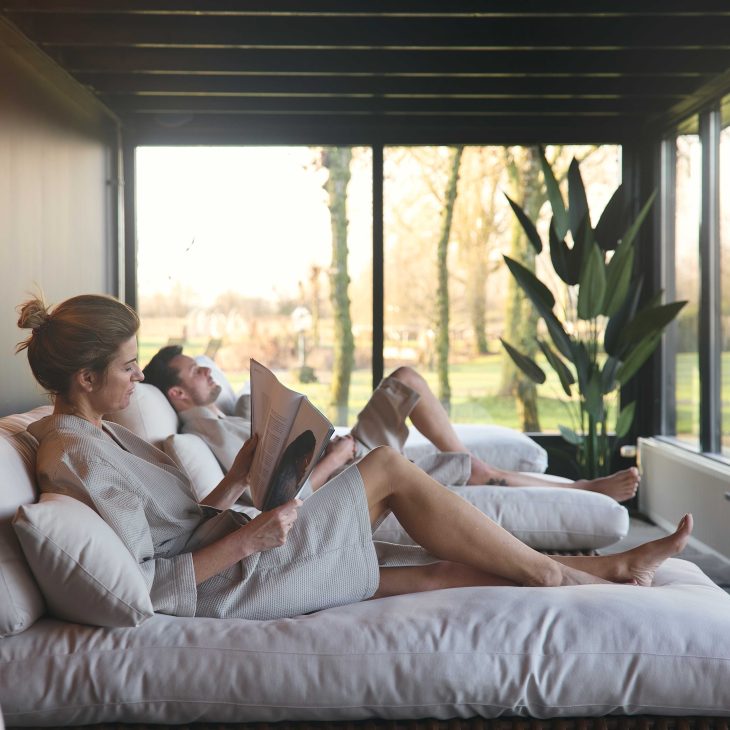Vrouw ligt relaxt een boek te lezen op een chill bed met een lichte badjas aan, op de achtergrond een gras weide met zonlicht.