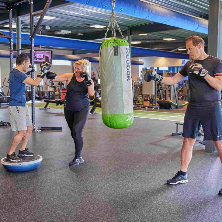 Verschillende mensen van verschillende leeftijden zijn aan het boksen: een man stoot tegen een bokszak, terwijl een vrouw bokst tegen een man met bokshandschoenen die op een balanceerbal staat.