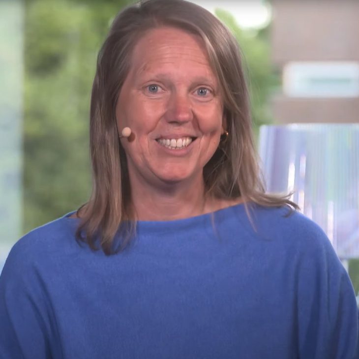 Een vrouw met een blauwe trui aan kijkt recht in de camera en heeft een glimlach, zij draagt een microfoon.