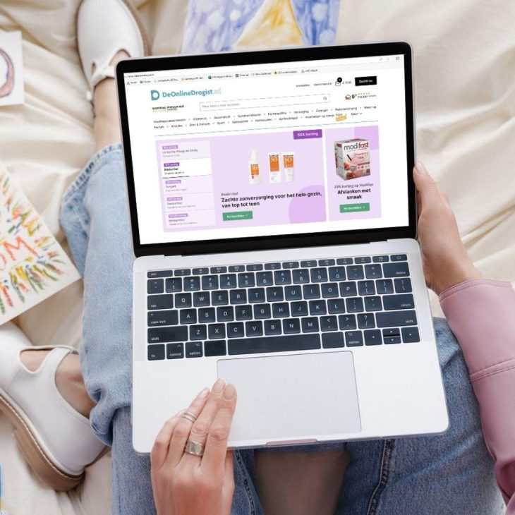 Een vrouw met een laptop op schoot die een webpagina open heeft staan van De Online Drogist. Op de pagina zijn verschillende zonnebrand aanbiedingen te zien.