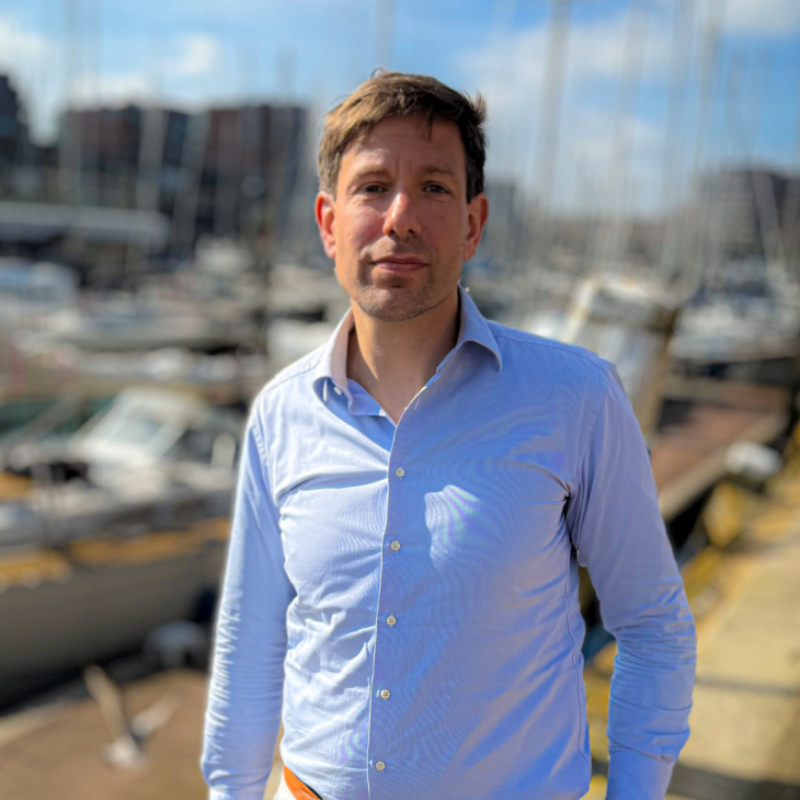 Een man in een blouse staat buiten in de zon, met op de achtergrond zeilboten in een haven.