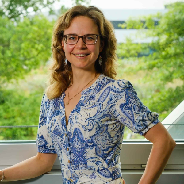 Anneke Valk, met lichtbruin haar, een blauw-witte jurk en een bril, lacht terwijl ze met haar hand in haar zij staat.