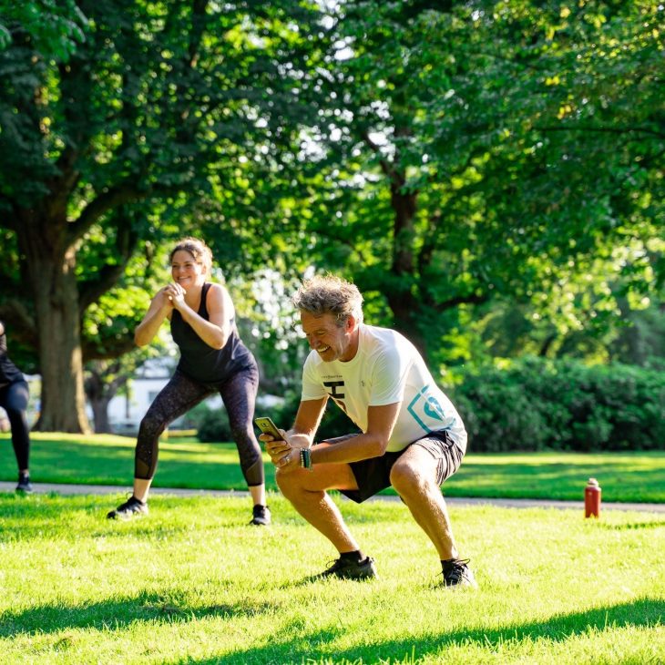 In een zonnig park doet een man een squat, terwijl op de achtergrond twee andere mensen ook aan het squatten zijn. Het gras is groen en de lente is duidelijk voelbaar.