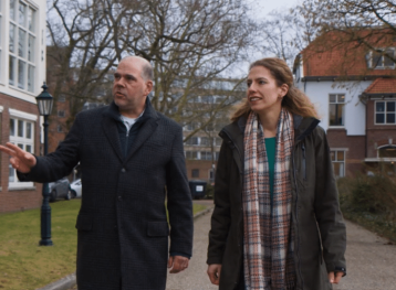 Still van video waarbij man en vrouw naast elkaar op straat lopen. Man draagt donkere jas, vrouw geruite sjaal en bruine jas