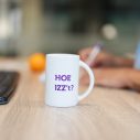 Op een bureau staat een koffiebeker met de tekst 'Hoe IZZ’t', met een sinaasappel en een toetsenbord op de achtergrond.