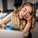 Een vrouw met blond haar zit achter haar computer en houdt vermoeid haar hand voor haar ogen.
