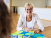 Vrouw met bril en krullend haar lacht terwijl ze aan tafel zit met stapels gekleurde doosjes.