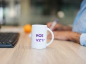 Op een bureau staat een koffiebeker met de tekst 'Hoe IZZ’t', met een sinaasappel en een toetsenbord op de achtergrond.