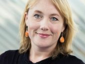 Vrouw met blond haar en oranje oorbellen kijkt glimlachend in de camera.
