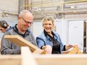 Blonde vrouw in spijkerjas lacht naar man in werkplaats